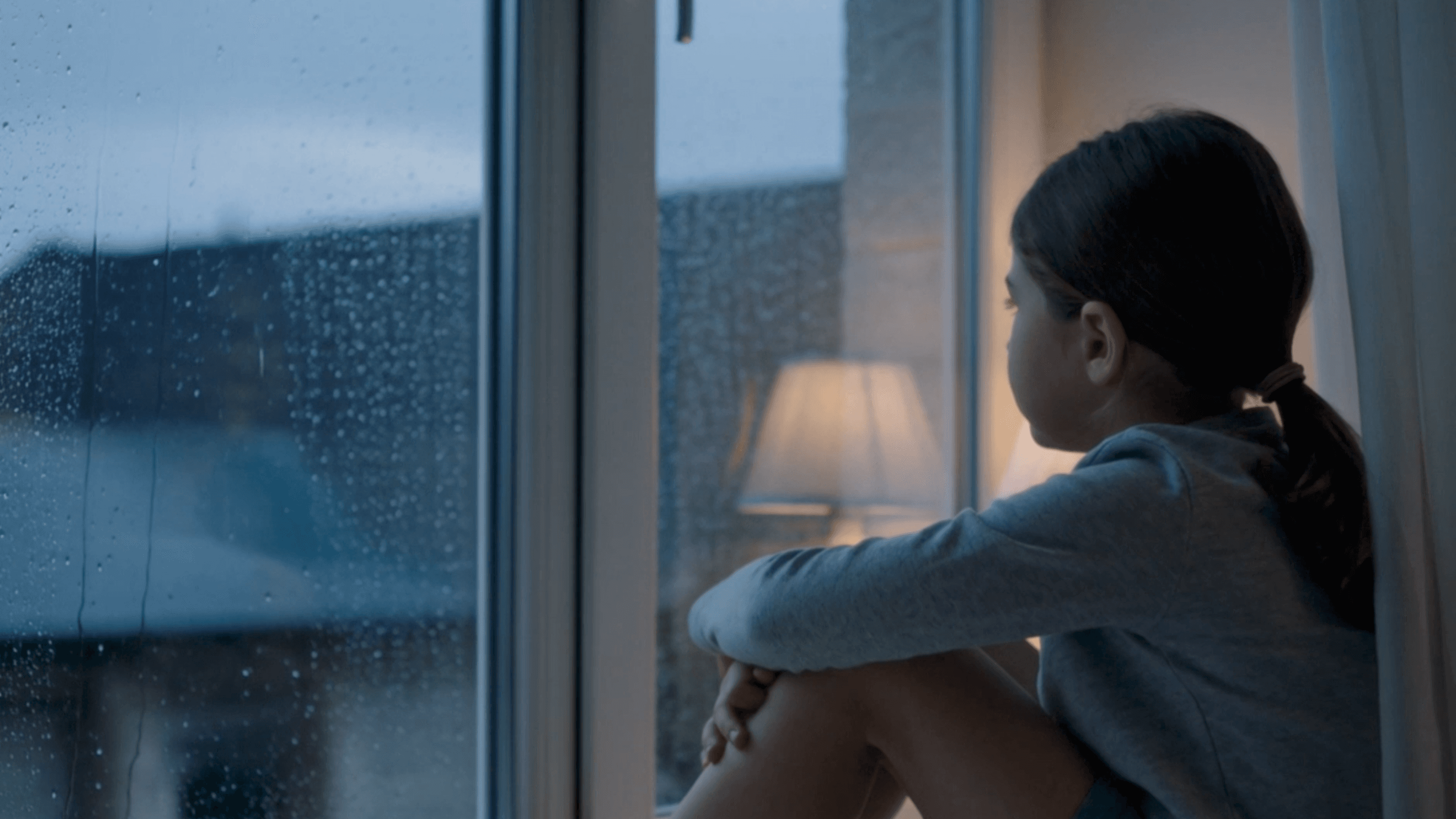 A young person sitting by a rain-streaked window at dusk, looking out thoughtfully.